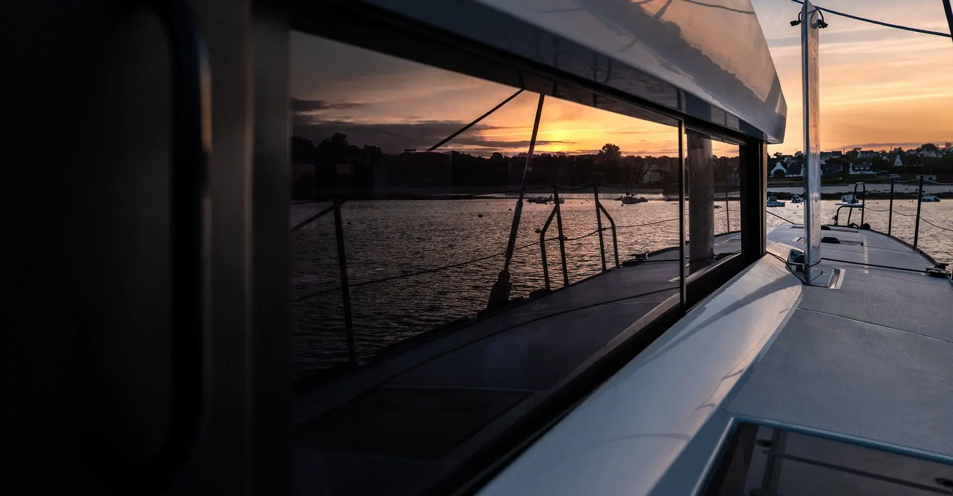 Sunset view along the deck of Nyota, a Windelo 50 electric performance catamaran, with the water reflected in the cabin windows.