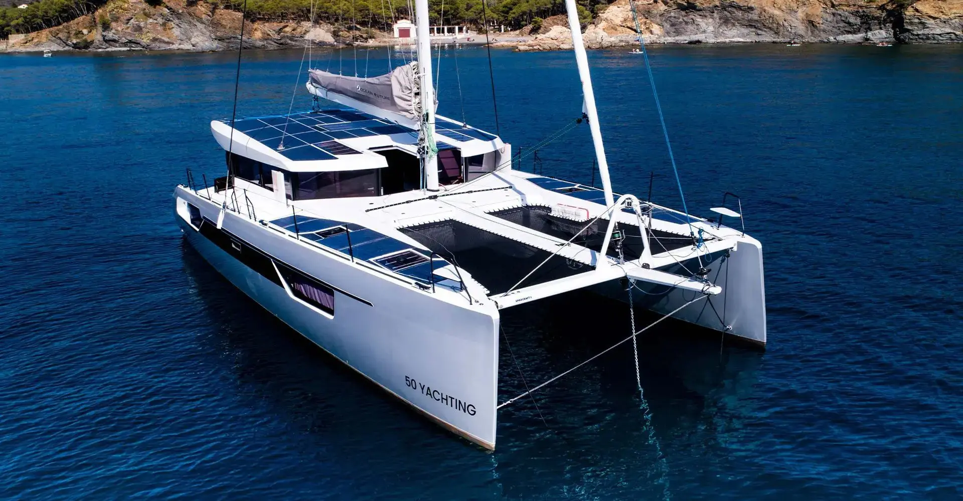Electric catamaran with deck solar panels anchored in a calm Mediterranean cove below a pine-covered rocky coast