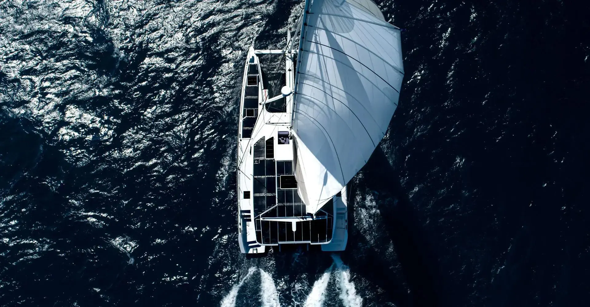 Overhead bird's-eye view of an electric catamaran sailing downwind under a large sail, solar panels on deck, twin wakes astern