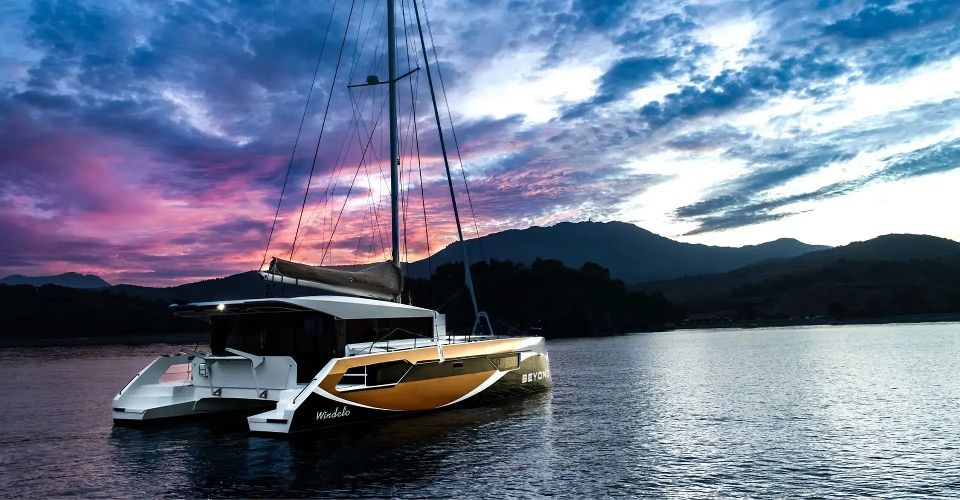 Electric performance catamaran anchored in a calm bay at dusk, cabins lit under a pink sunset sky