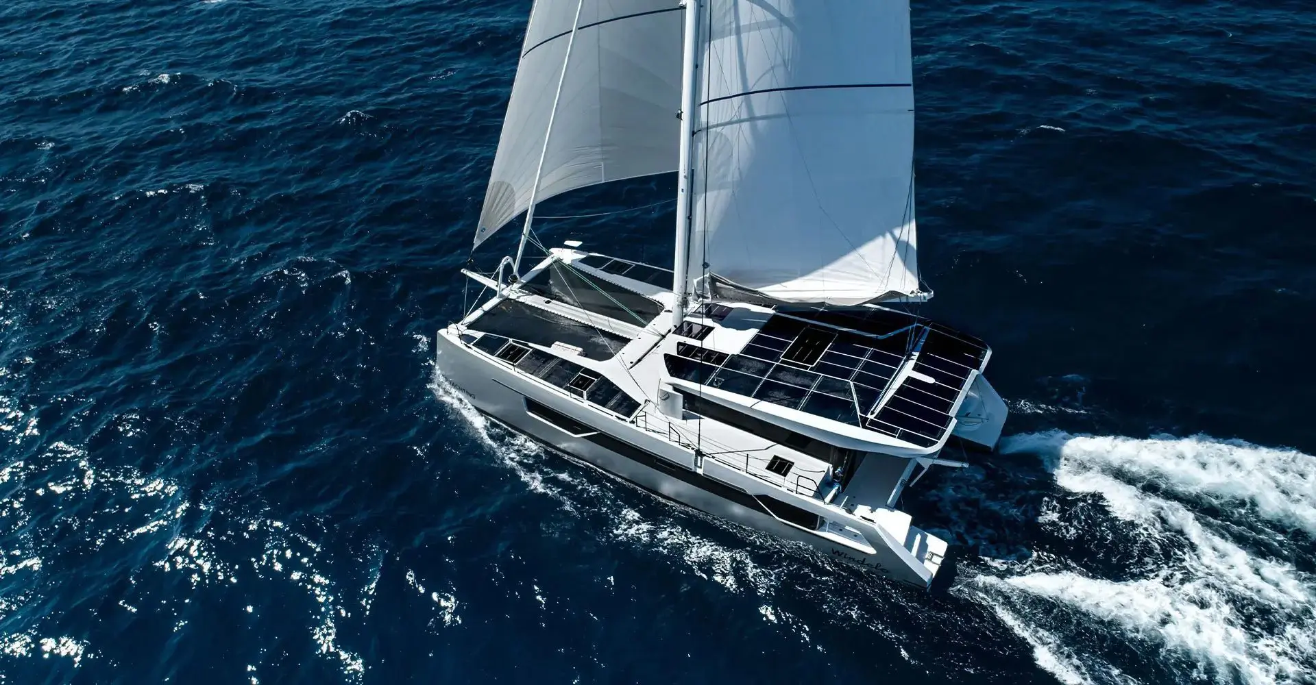 Electric performance catamaran sailing under full main and headsail, solar panels on the hardtop, aerial view