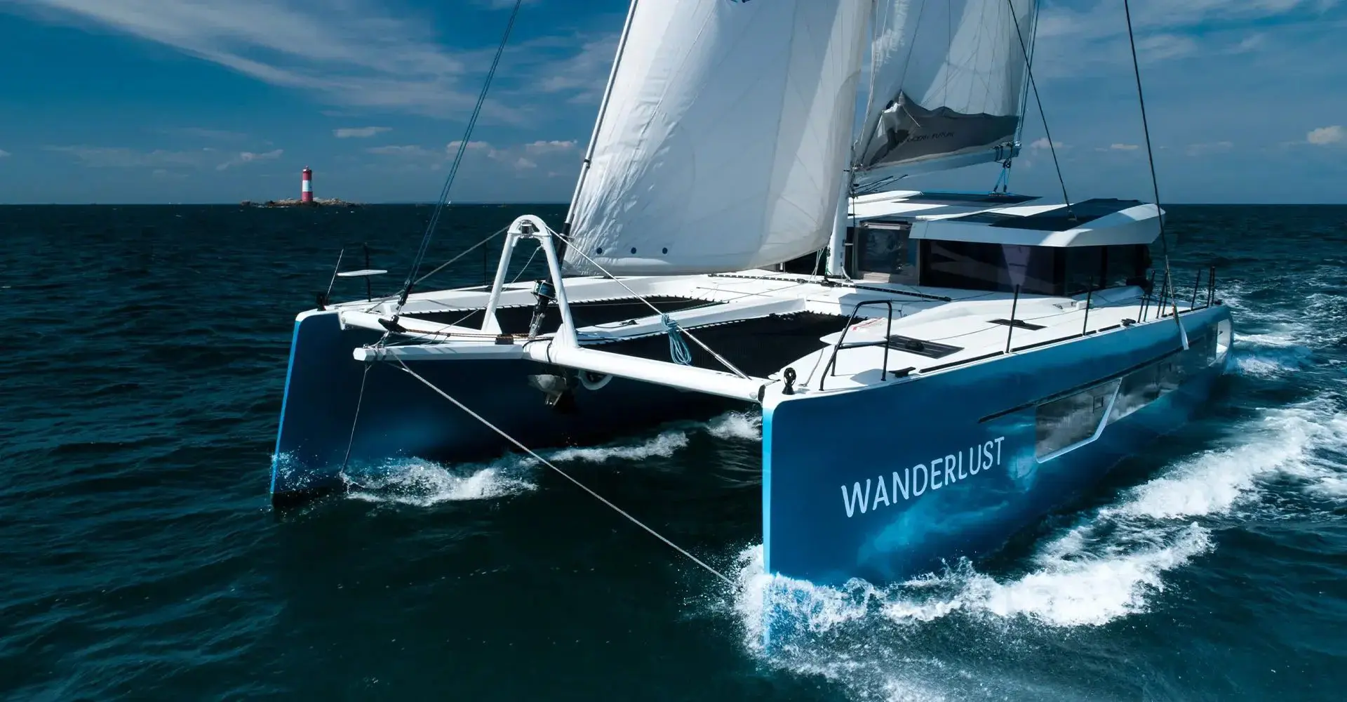 Blue-hulled electric performance catamaran sailing through open water with spray at the bows, lighthouse on the horizon