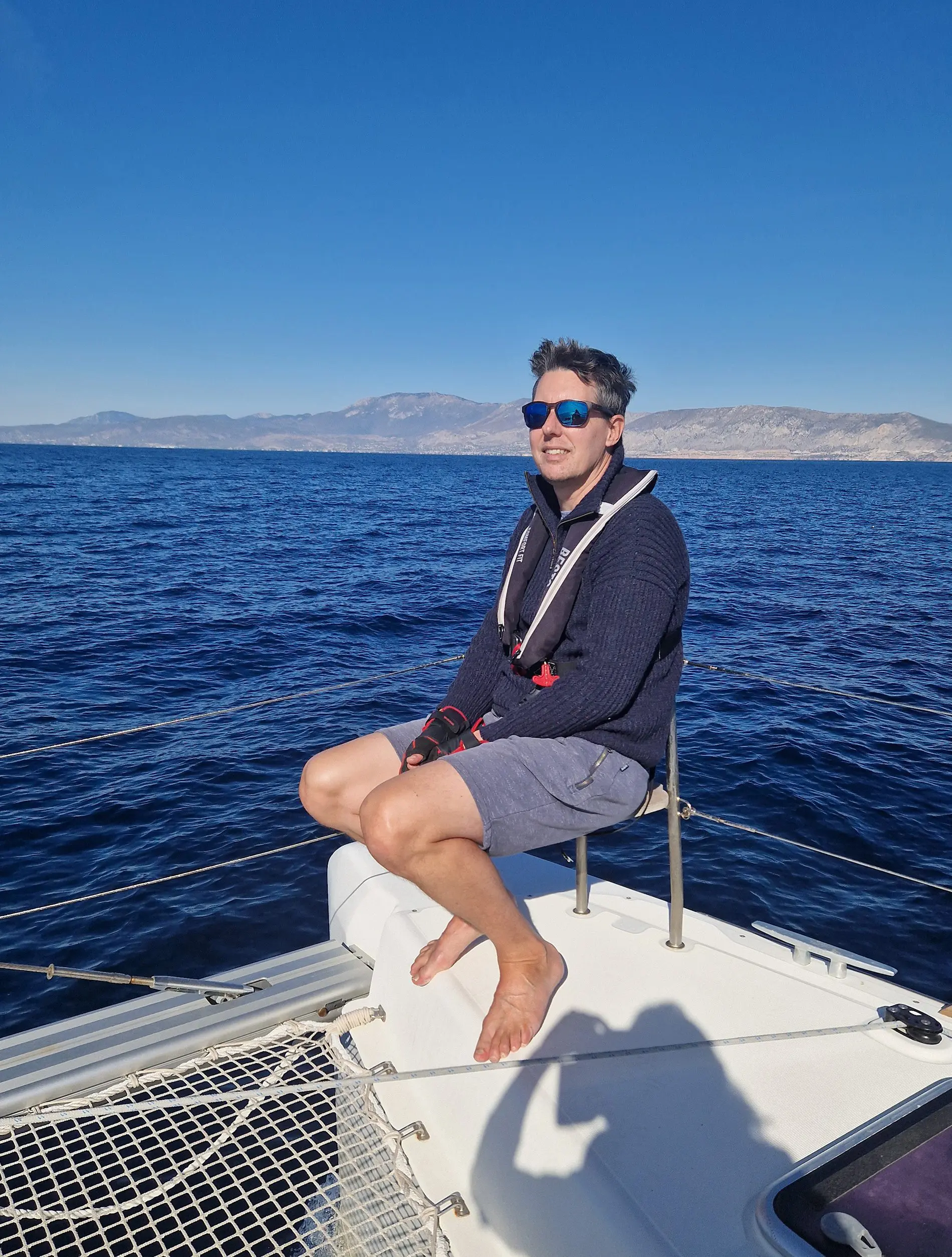 Joop Marijne, owner and coach of Volt Sail, seated on the bow of a catamaran with open sea and mountains behind
